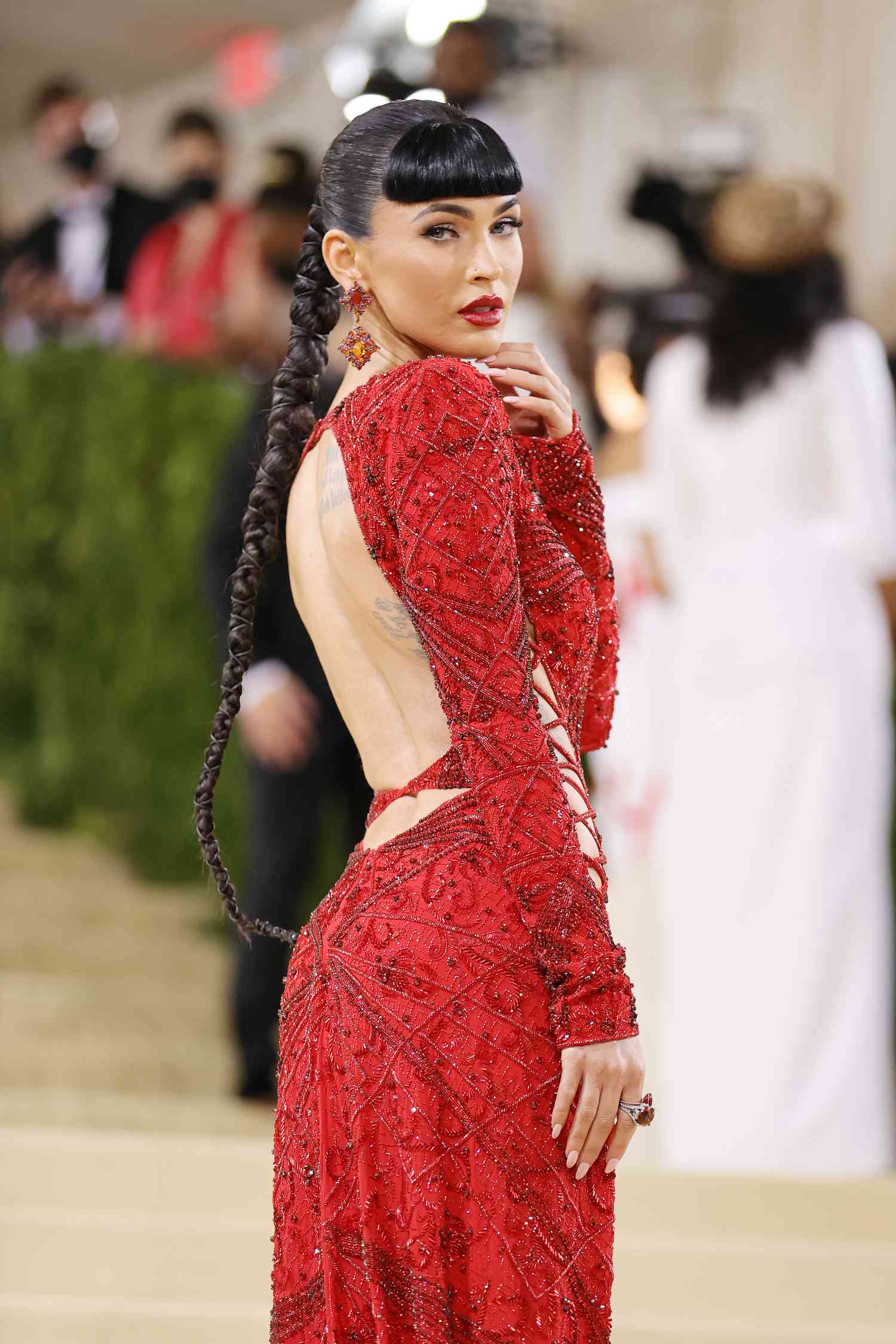 Megan Fox at the Met Gala in a backless red dress with blunt black bangs and a long braided ponytail