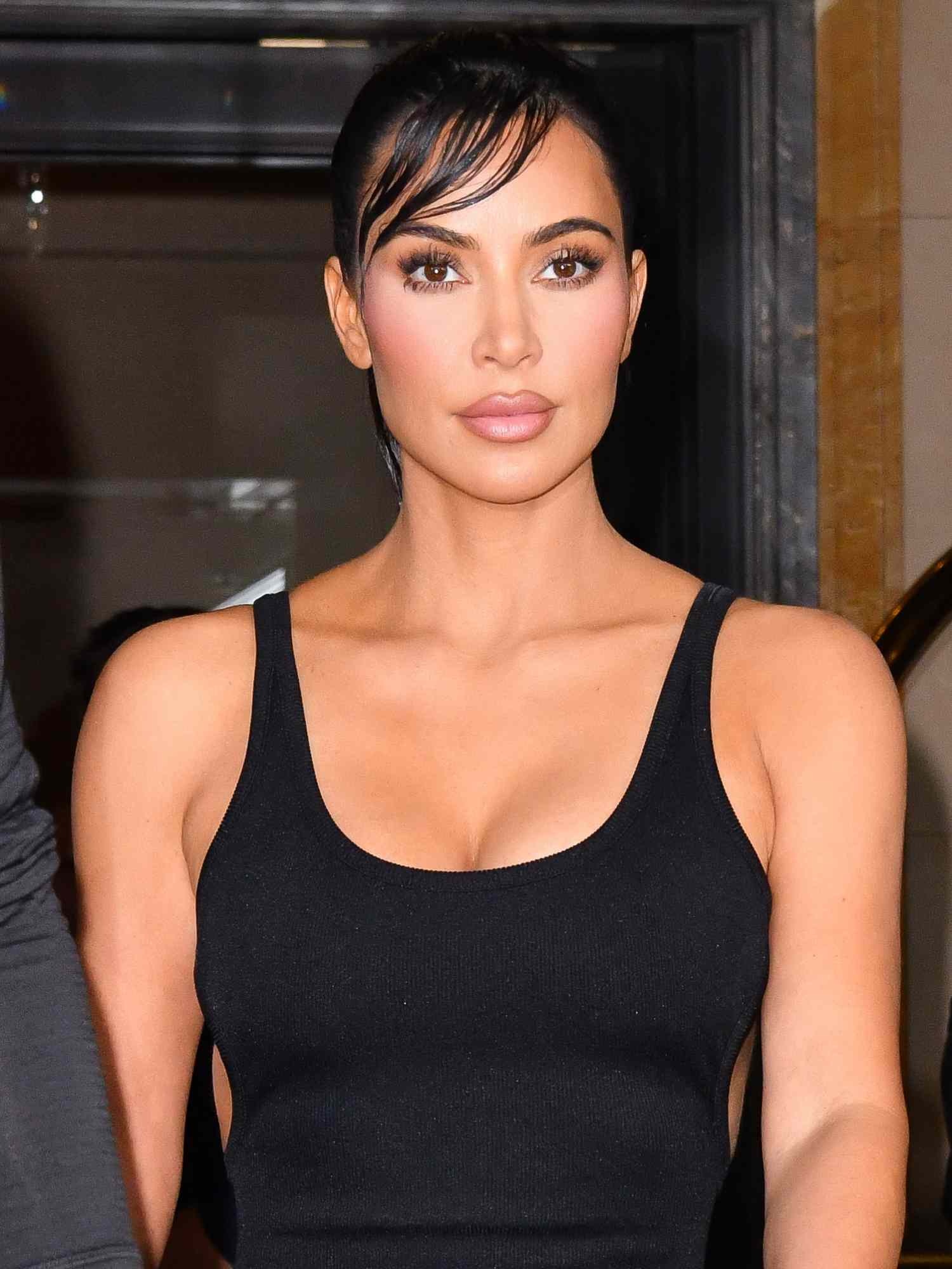 Kim Kardashian wears a black tank top, slicked back hair with side bangs, and blush-toned makeup look