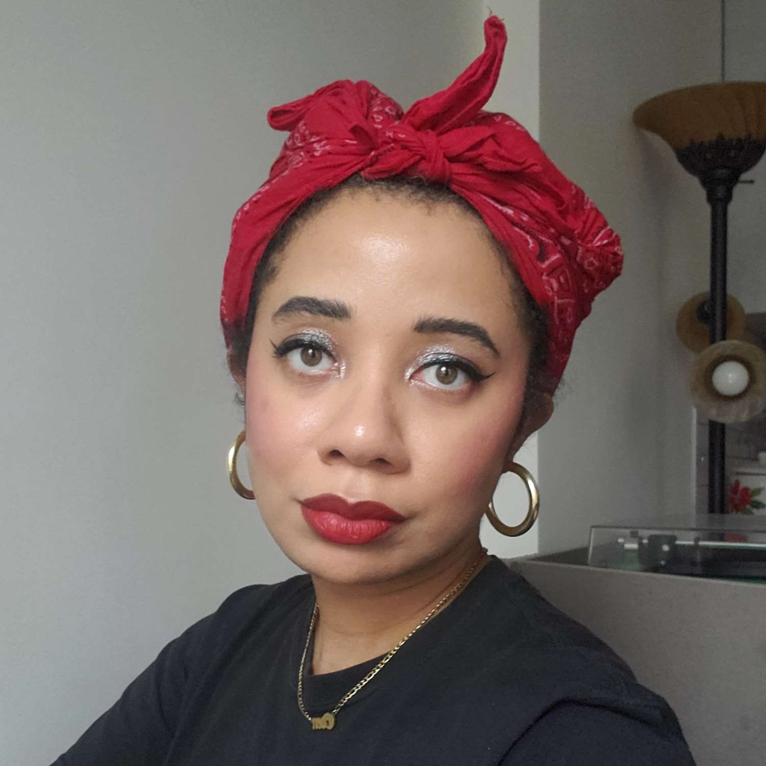 Byrdie editor Eden Stuart wears a glittery eye makeup look with winged liner, blush, red lipstick, gold hoop earrings, and a red bandana