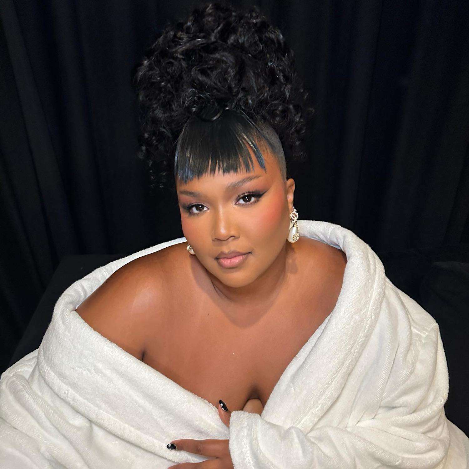 Lizzo wears a voluminous curly updo hairstyle with baby bangs and pearl drop earrings