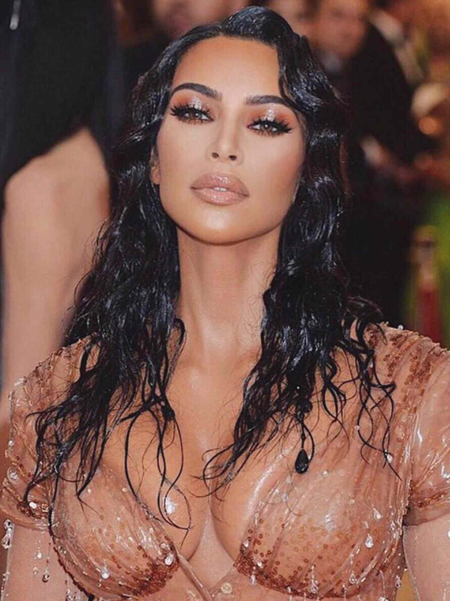 Kim Kardashian wears a nude dress, wet look wavy hair, and makeup look with orange/bronze eyeshadow, mascara, and glossy nude lip