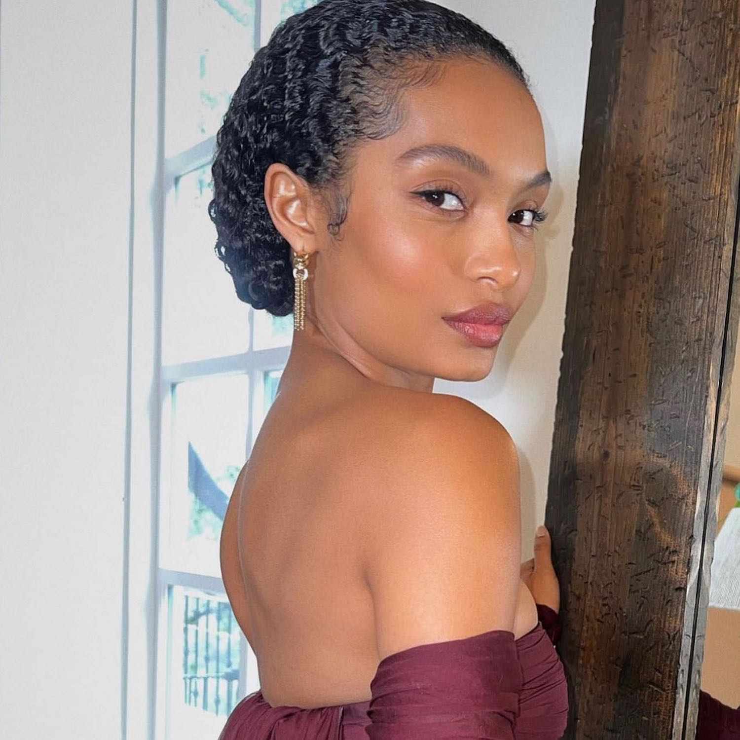 Yara Shahidi wears a slicked-back low curly updo and radiant makeup