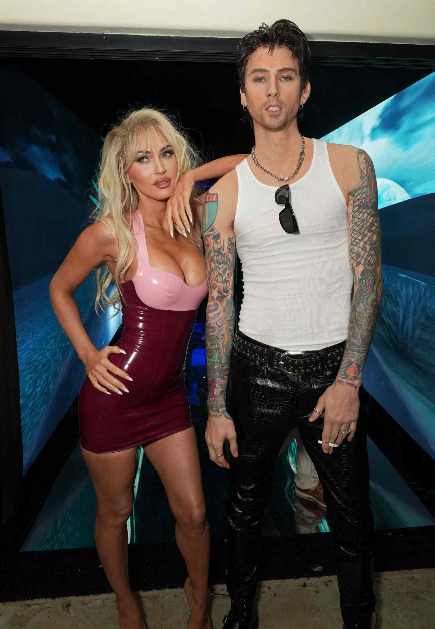 Megan Fox and Machine Gun Kelly at the Casamigos Halloween Party in 2022 dressed up as Pamela Anderson and Tommy Lee. Fox is in a blonde wig and a pink and red mini dress and MGK is in a white tank top and black leather pants with black hair.