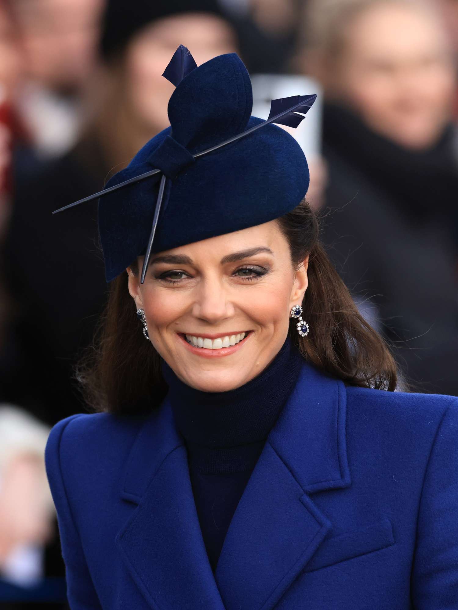 Kate Middleton in a blue coat, black turtleneck sweater, blue hat, and fresh makeup look