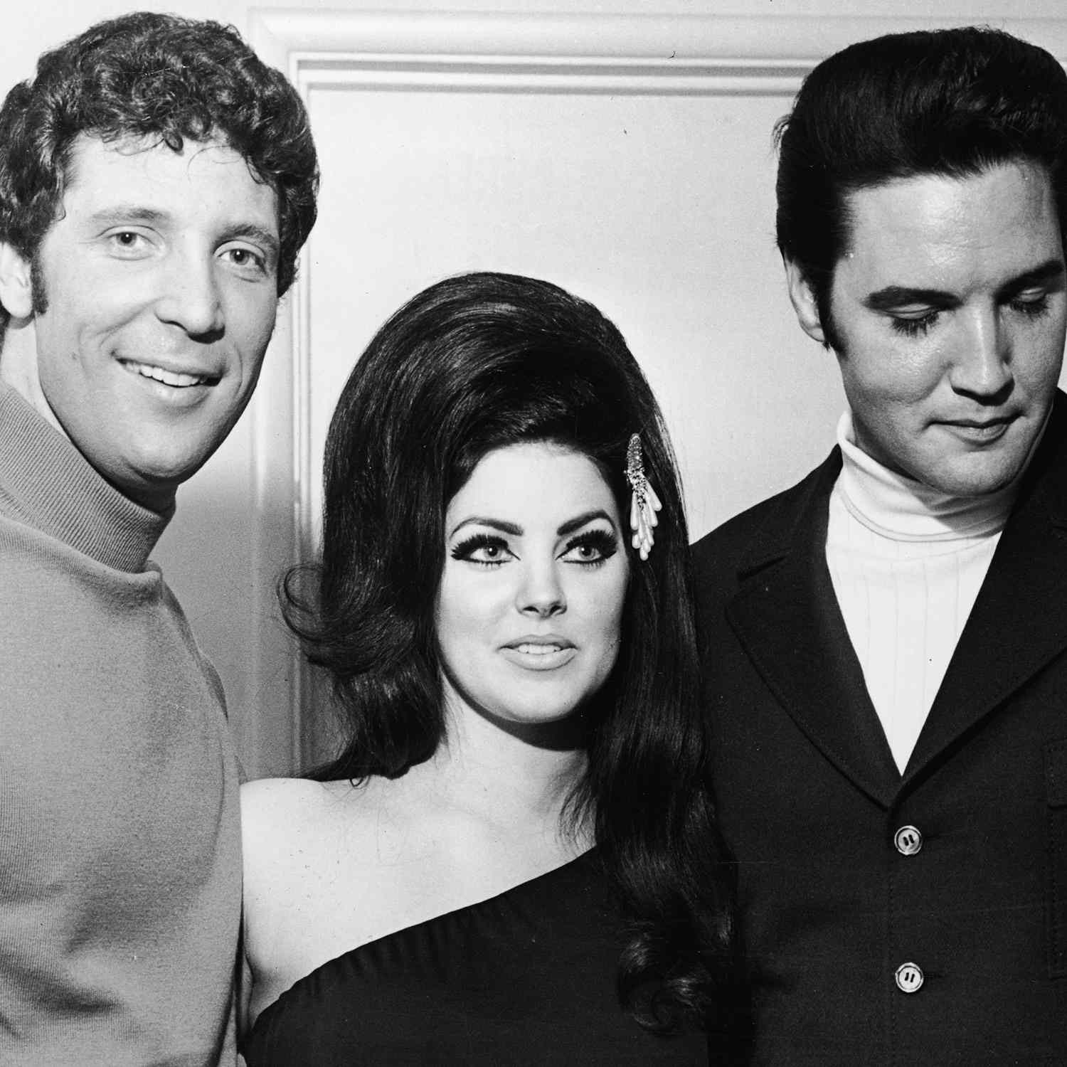 Priscilla Presley next to Elvis Presley and a friend. Priscilla has a huge side sweeping bump in her hair with an off the shoulder dress and her iconic eyeliner