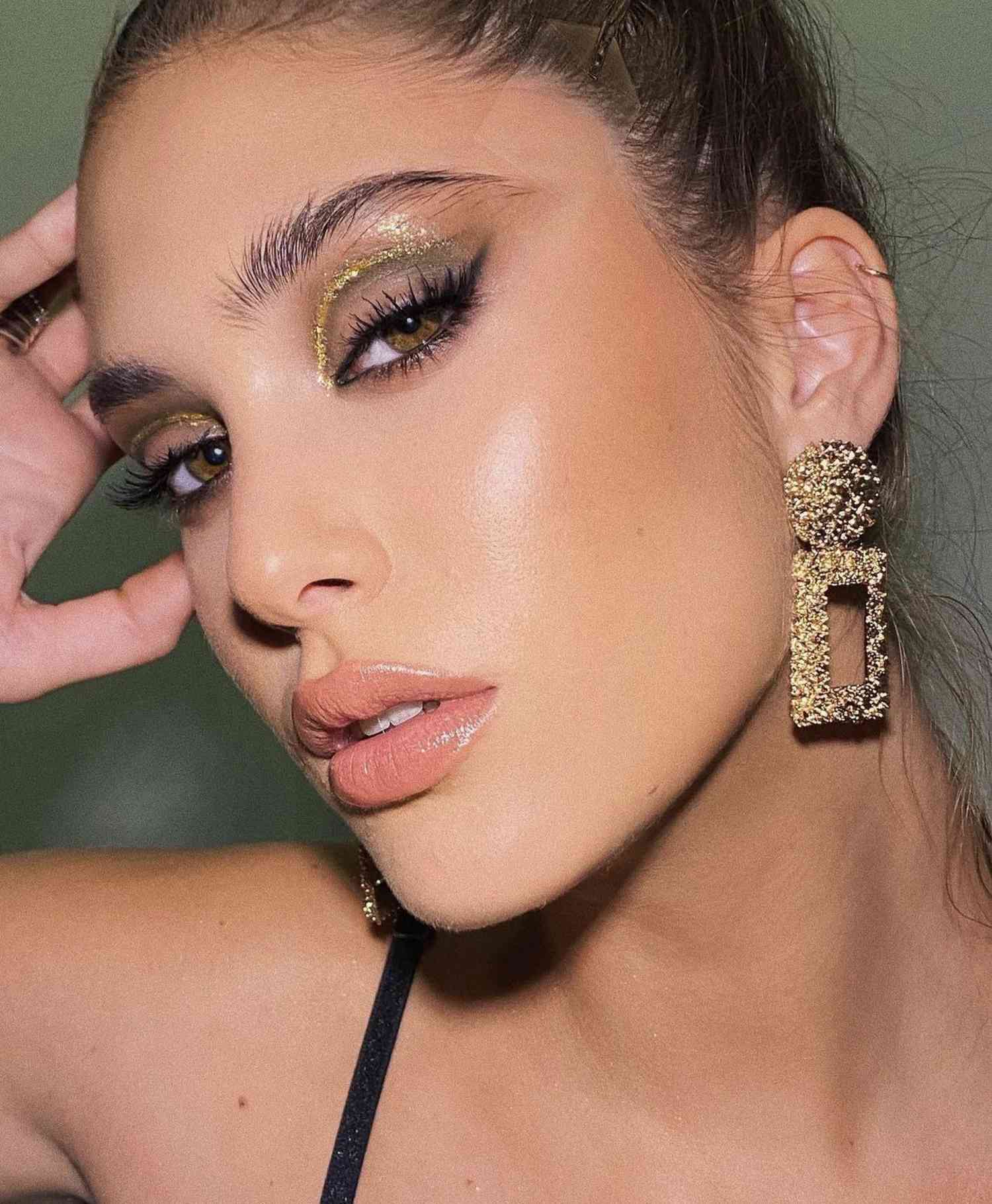 woman wearing gold glitter in the crease of the lid with black wing liner and false lashes paired with a light pink nude liptick