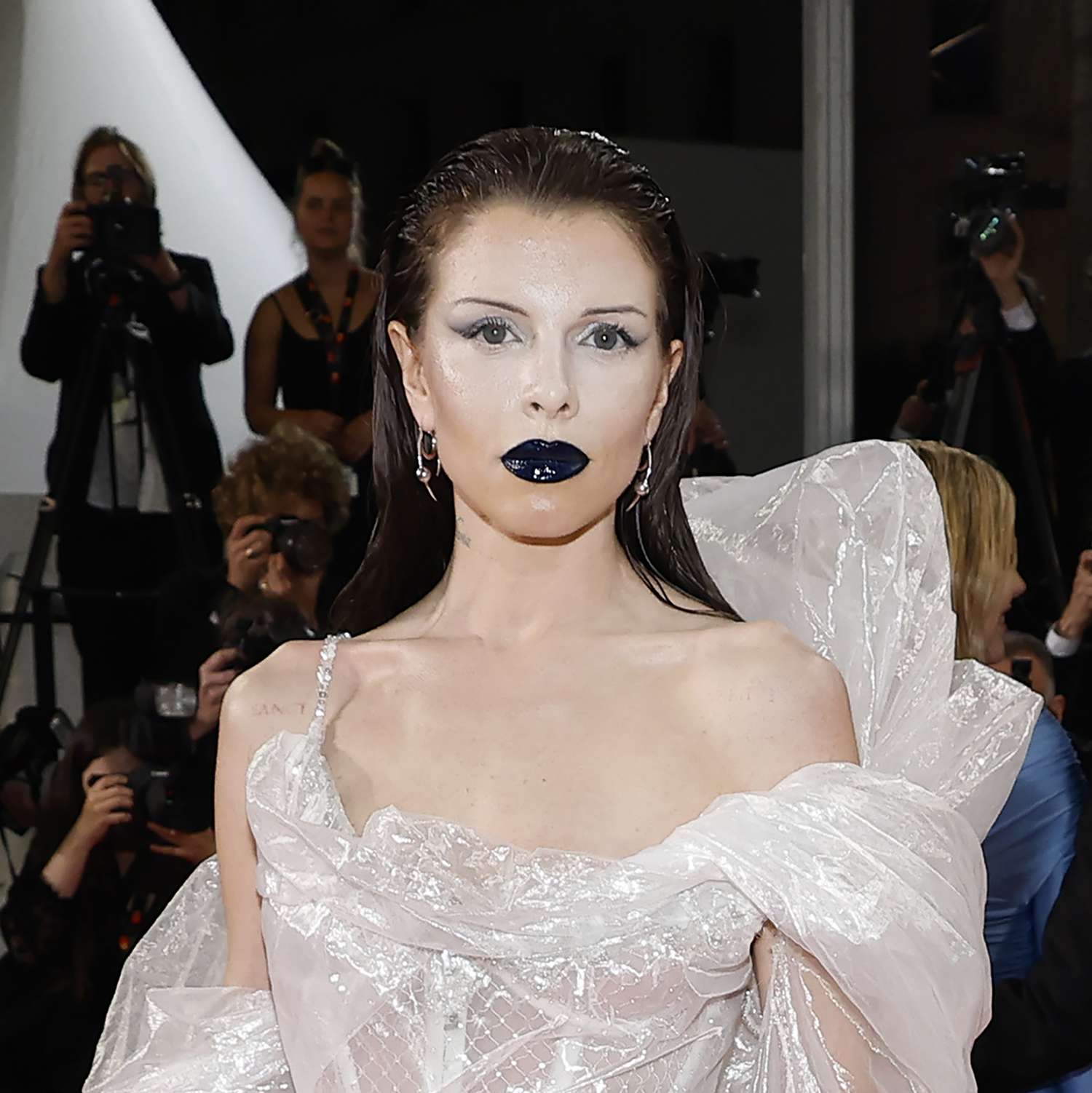 Julia Fox is seen at Cannes wearing white airbrushed makeup on her face, with a tan contour around it around the jawline, and black lipstick with gloss on top, and thin eyebrows. Her eye makeup is a soft shadow wing extended in the outer corners 