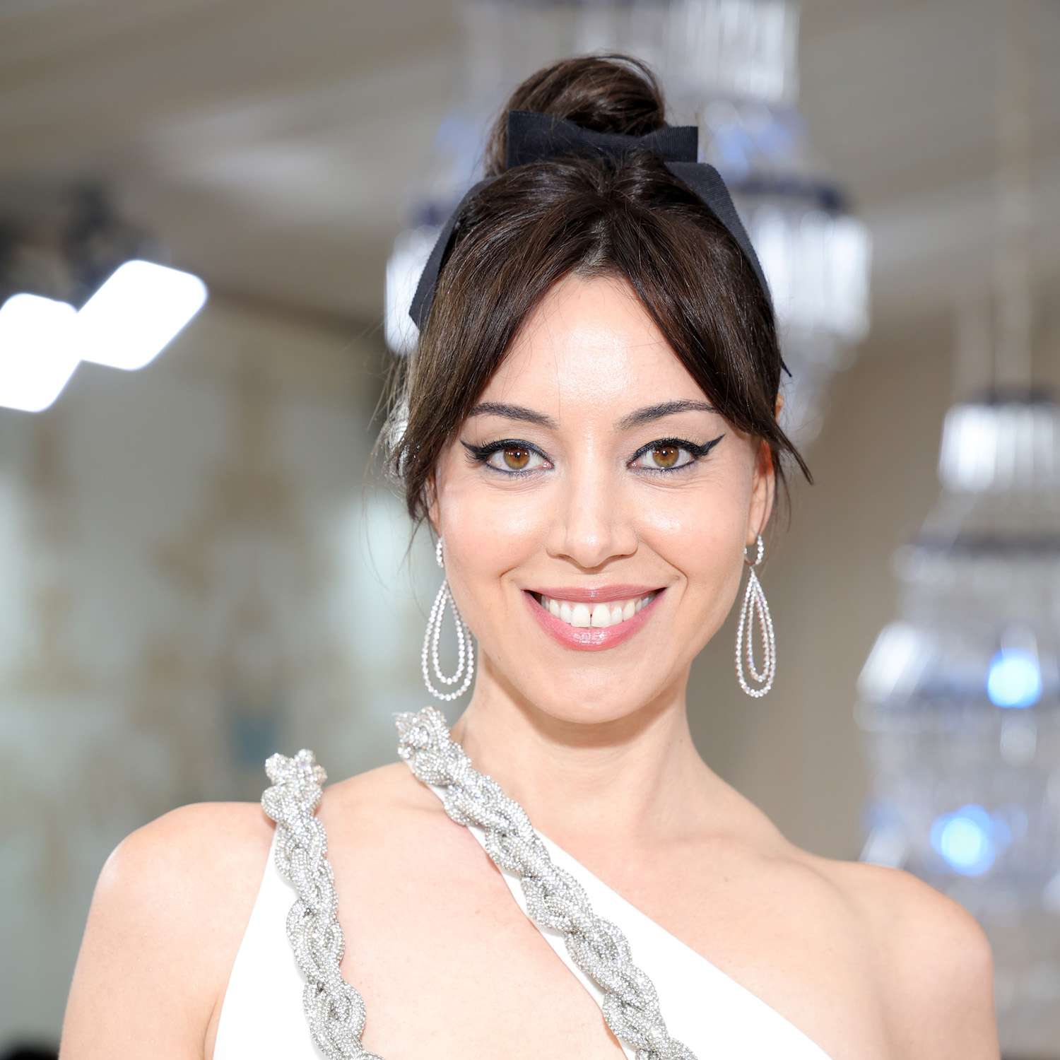 Aubrey Plaza wears a high bun with black ribbon and curtain bangs and a white Stella McCartney dress to the 2023 Met Gala