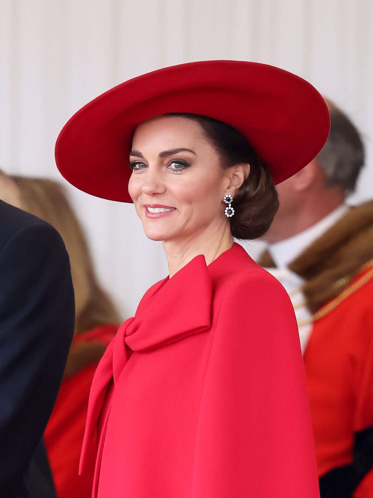 Kate Middleton in a red cape and red hat with an updo hairstyle and fresh makeup look