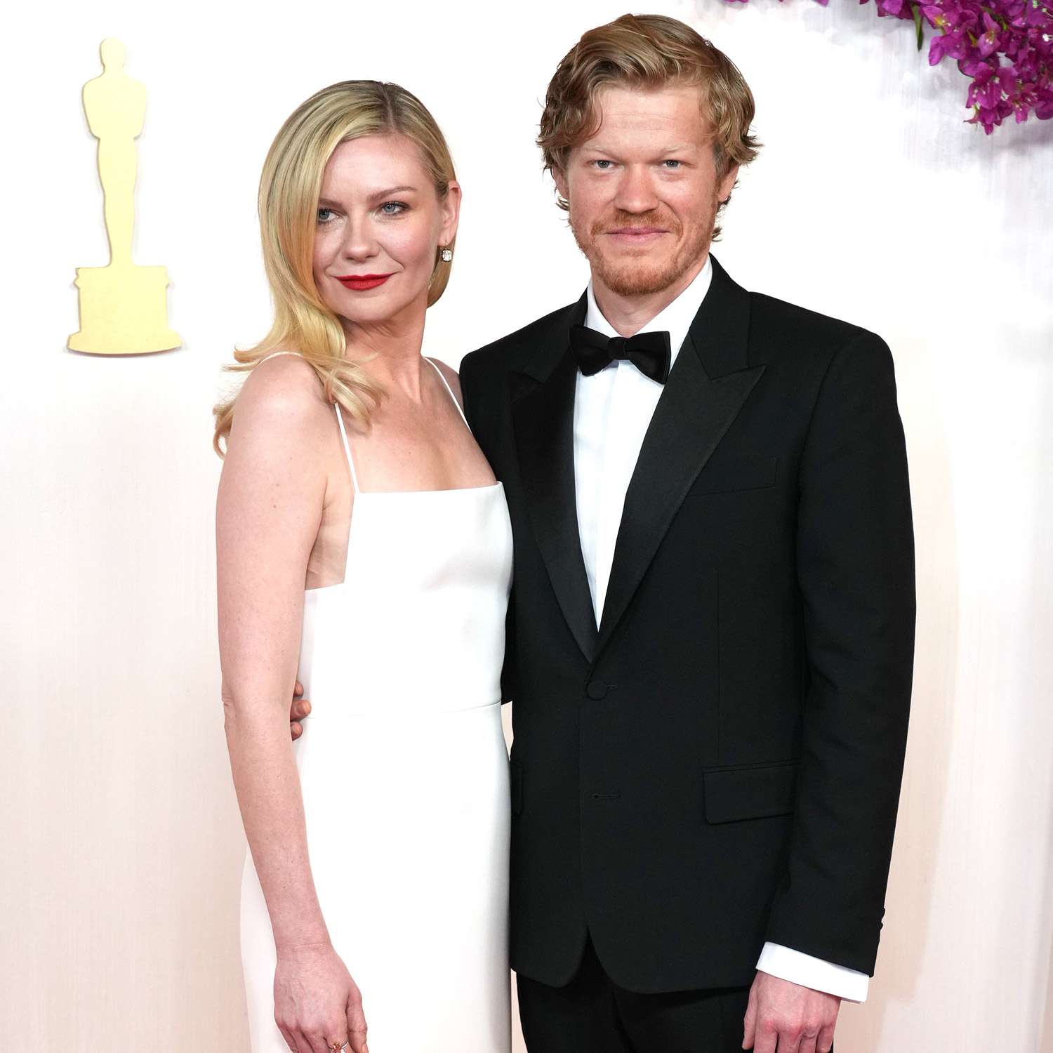 Kirsten Dunst on the 2024 oscars red carpet with blonde hair, a white Gucci dress, and bright red lipstick standing next to her husband Jesse plemons