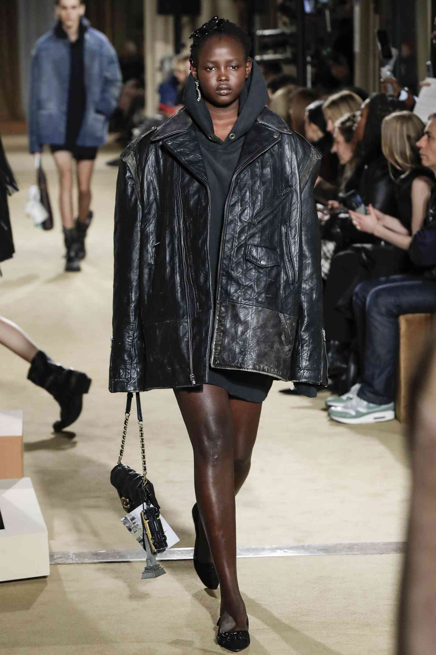 A model walks the Coach runway wearing a black hoodie with a black oversized leather biker jacket. 