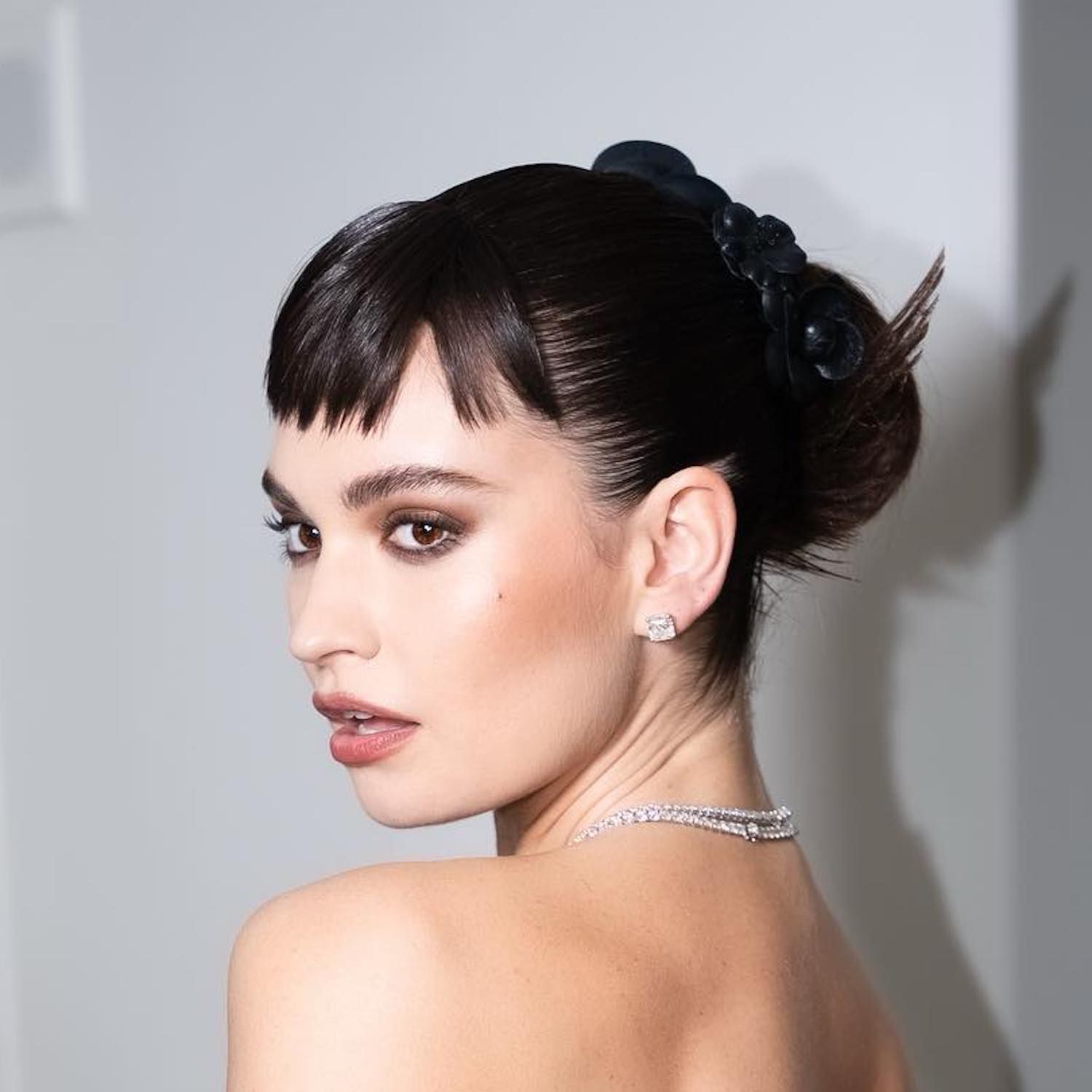 Lily James wears a spiky bun hairstyle with baby bangs and black camellia hair accessories