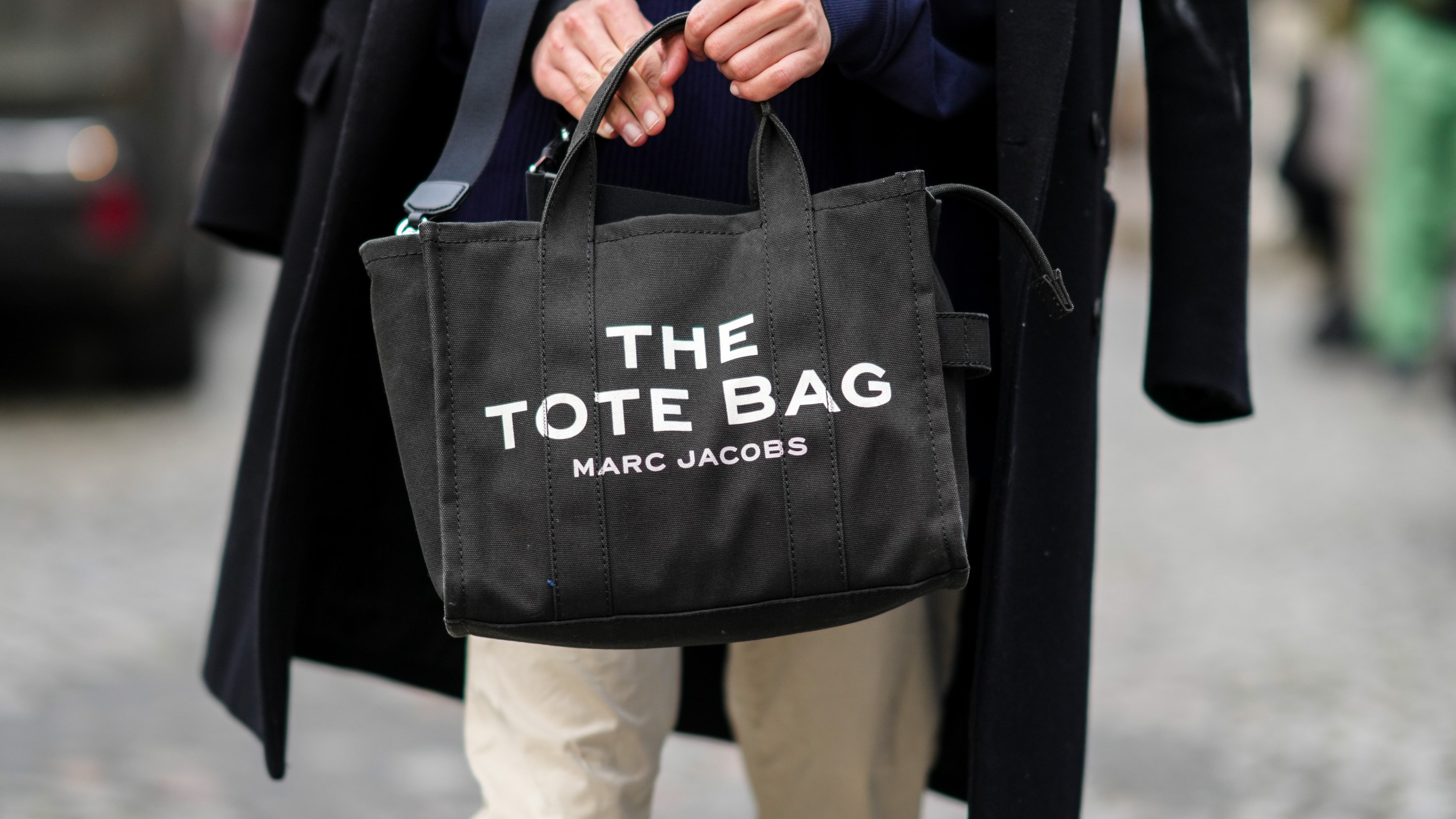 A guest wears a navy blue ribbed wool pullover, a black long coat, a black Tote-bag handbag from Marc Jacobs, beige pants, outside Hermes, during Paris Fashion Week - Menswear F/W 2022-2023, on January 22, 2022 in Paris, France.