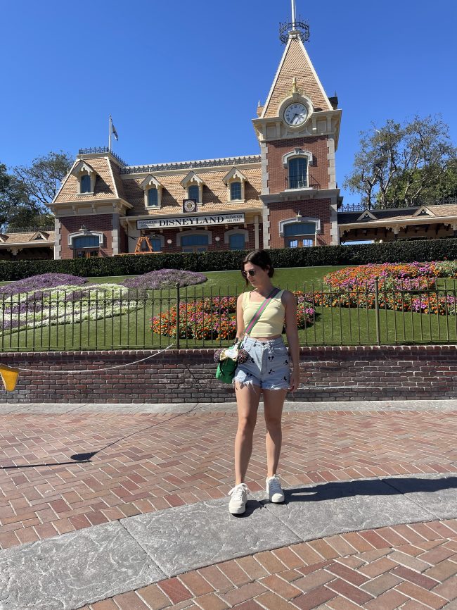 A woman wearing white sneakers, jean shorts with Mickey heads embroidered on them, and a yellow tank top, standing in front of the Disneyland entrance. 