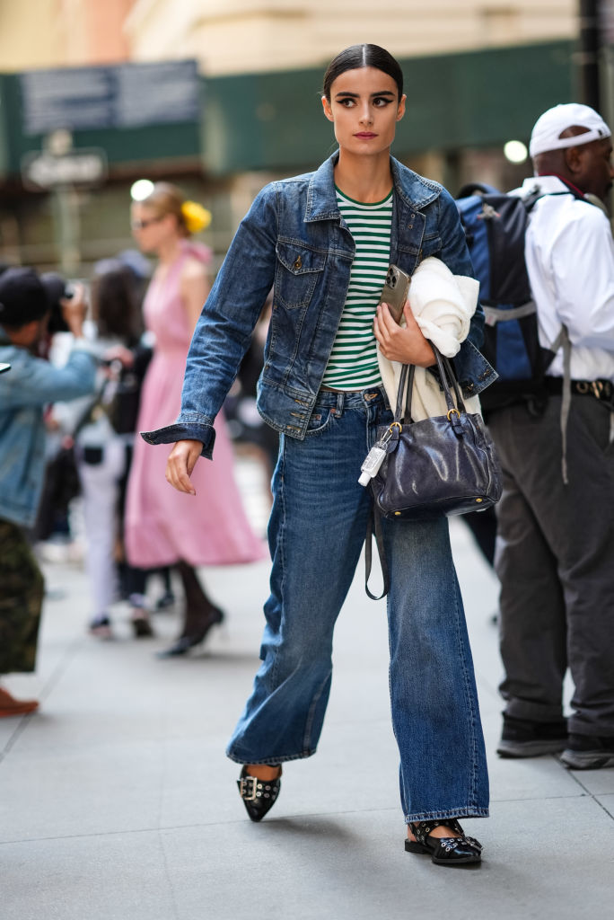 NEW YORK, NEW YORK - SEPTEMBER 09: A model wears white and dark green striped shirt, navy denim jean jacket, navy denim jean high waisted pants, black pointed toe belted Ganni ballerina leather shoes, dark gray leather bag, white jacket in hand, outside Carolina Herrera, during the New York Fashion week Spring/Summer 2025 on September 9, 2024 in New York, New York.