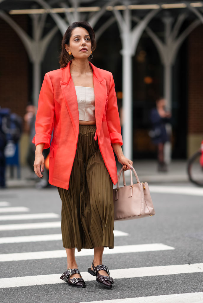 NEW YORK, NEW YORK - SEPTEMBER 07: A guest wears orange earrings, cream cropped top, bright orange red blazer jacket, dark green pleated midi skirt, light brown leather bag, shiny black belted Ganni belted ballerina flats leather shoes , outside Tibi, during the New York Fashion week Spring/Summer 2025 on September 07, 2024 in New York, New York.