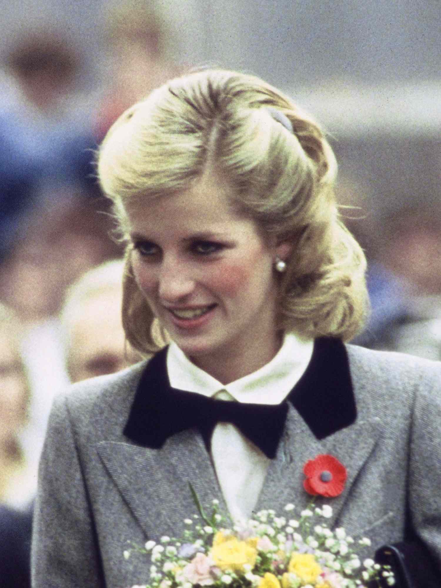 Princess Diana wears her hair pinned back, a gray blazer, and a white blouse with black bow tie