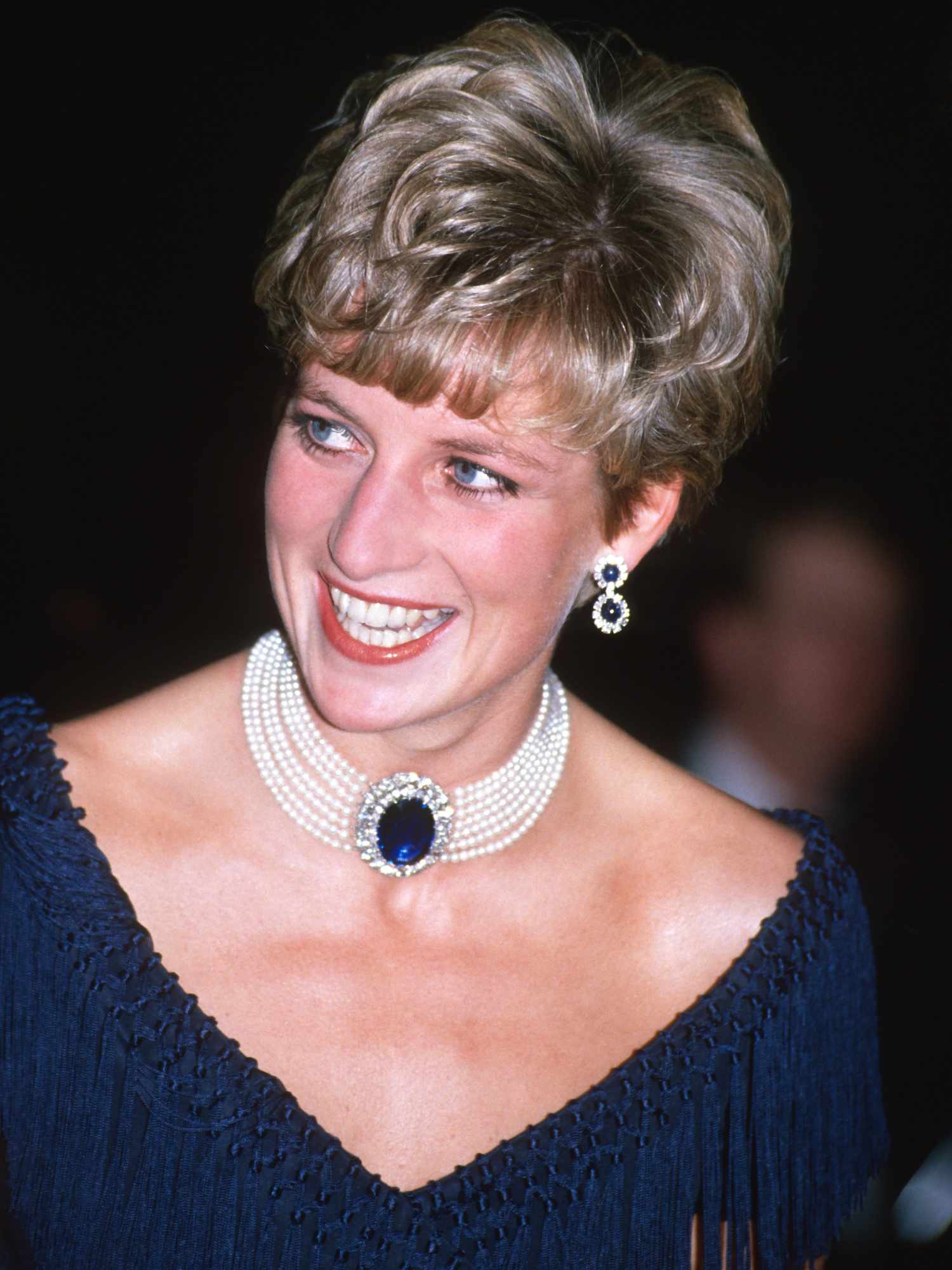 Princess Diana wears a short voluminous pixie haircut with bangs, sapphire jewelry, and a blue fringed cocktail dress